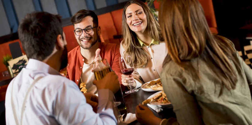Grupo de amigos disfrutando de una cena en un restaurante, riendo y compartiendo bebidas y comida en un ambiente acogedor.